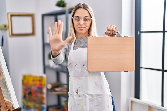 Young blonde painter woman holding wooden case at art studio with open hand doing stop sign with serious and confident expression, defense gesture
