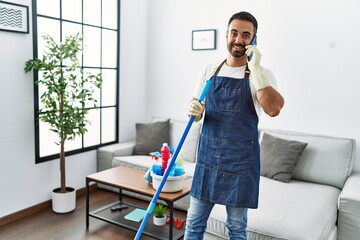 Young hispanic man wearing apron and gloves talking on the smartphone at home