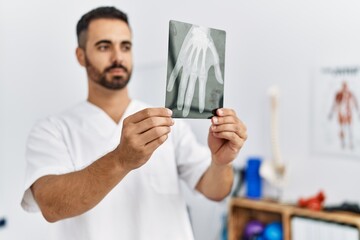 Young hispanic man wearing physiotherapist uniform holding hand xray at clinic