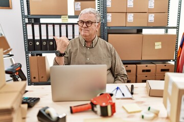 Senior caucasian man working at small business ecommerce with laptop smiling with happy face looking and pointing to the side with thumb up.