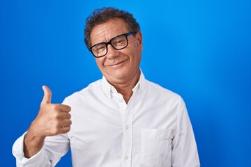 Middle age hispanic man standing over blue background doing happy thumbs up gesture with hand. approving expression looking at the camera showing success.