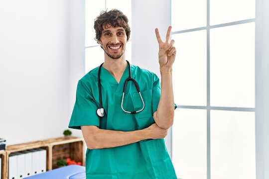 Young hispanic man wearing doctor uniform and stethoscope at clinic smiling with happy face winking at the camera doing victory sign. number two.