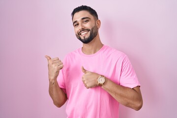 Hispanic young man standing over pink background pointing to the back behind with hand and thumbs...