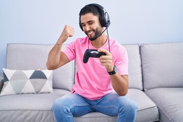 Hispanic young man playing video game holding controller sitting on the sofa very happy and excited doing winner gesture with arms raised, smiling and screaming for success. celebration concept. © Krakenimages.com