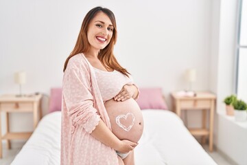 Young pregnant woman touching belly with heart lotion symbol standing at bedroom