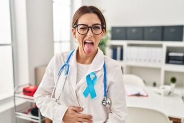 Young brunette doctor woman wearing stethoscope at the clinic sticking tongue out happy with funny...