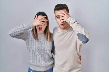 Young hispanic couple standing over white background peeking in shock covering face and eyes with hand, looking through fingers with embarrassed expression.