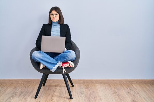 Young Hispanic Woman Sitting On Chair Using Computer Laptop Skeptic And Nervous, Frowning Upset Because Of Problem. Negative Person.