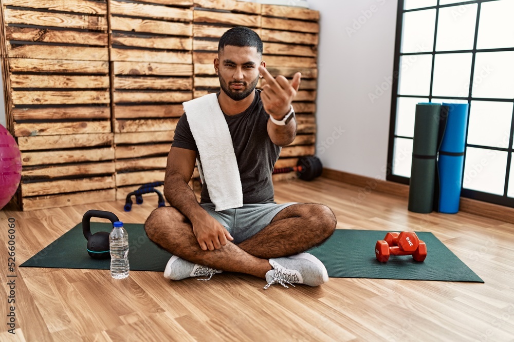 Poster young indian man sitting on training mat at the gym showing middle finger, impolite and rude fuck of