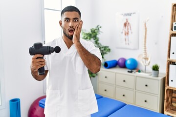 Young indian physiotherapist holding therapy massage gun at wellness center afraid and shocked,...