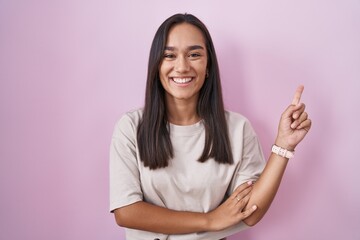 Young hispanic woman standing over pink background with a big smile on face, pointing with hand finger to the side looking at the camera.