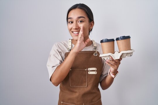 Young hispanic woman wearing professional waitress apron holding coffee hand on mouth telling secret rumor, whispering malicious talk conversation