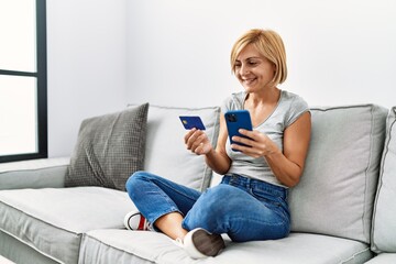 Middle age blonde woman smiling confident using smartphone and credit card at home