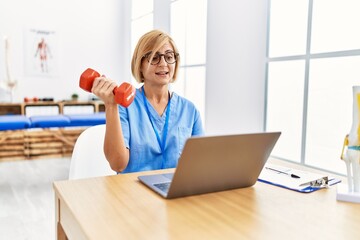Middle age blonde woman wearing physio therapy uniform doing tele rehab using dumbbells at clinic