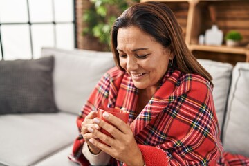 Middle age hispanic woman drinking coffee covering with blanket at home