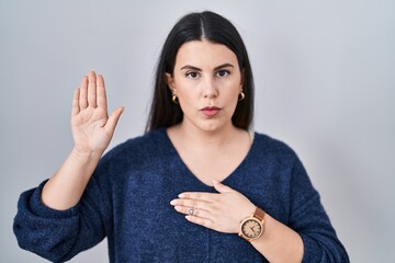 Young brunette woman standing over isolated background swearing with hand on chest and open palm,...