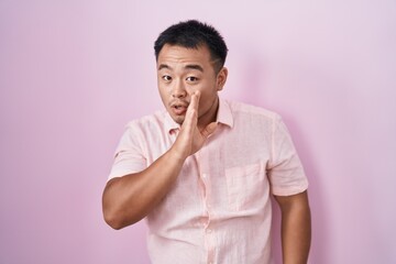 Chinese young man standing over pink background hand on mouth telling secret rumor, whispering malicious talk conversation