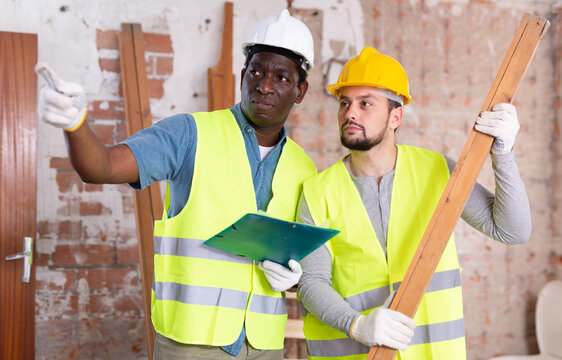 Builders discussing work in cosntruction site indoors. African-american man pointing finger, caucasian man holding wooden plank.