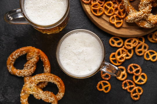 Mugs of fresh beer and pretzels on dark background, closeup. Oktoberfest celebration