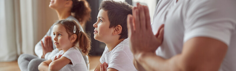 Fototapeta premium Calm woman is enjoying practice with husband and children and expressing namaste sign in lotus position