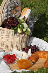 Picnic blanket with tasty food, flowers, basket and cider on green grass outdoors