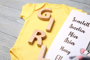 Bodysuit, list of baby names and toys on grey wooden background, closeup