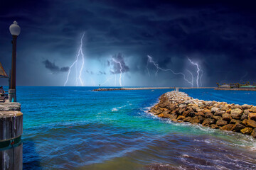 a gorgeous summer landscape at the beach with blue ocean water and a jetty made of large rocks with boats in the harbor and lush green palm trees and powerful storm clouds and lightning - Powered by Adobe