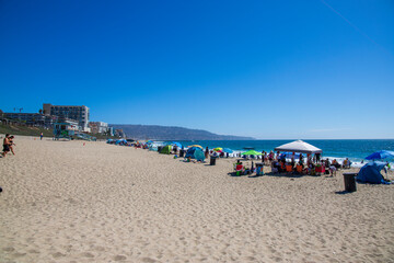 Fototapeta na wymiar a gorgeous summer landscape at the beach with blue ocean water and people relaxing in the brown sand under colorful umbrellas with a gorgeous clear blue sky at Redondo Beach Pier in Redondo Beach