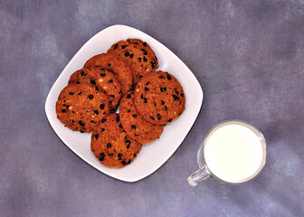 A plate with several oatmeal chocolate chip cookies and a glass of hot milk on a gray background.