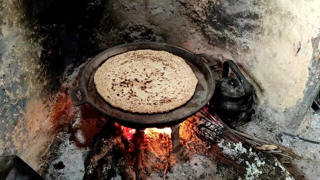 Close-up of Cooking bread in a cast iron skillet resting on trivets, over an oak wood fire in a large stone fireplace in a traditional kitchen. Medieval kitchen utensils. Morocco.