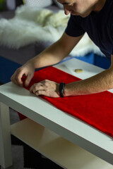 Naklejka premium Hands of experienced worker in the handmade industry, performing cutting tasks on red fabric, on a white surface, the beginning of red hat production. Homemade sewing. Working tailor. Christmas hat.