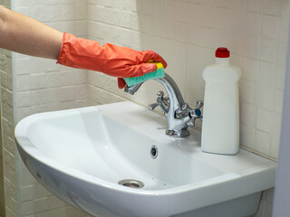 Cleaning bathroom sink and faucet with detergent in orange rubber gloves with green sponge. Housework, cleaning, hygiene home concept. Hand holds a mint sponge with white foam and scrubs the faucet.