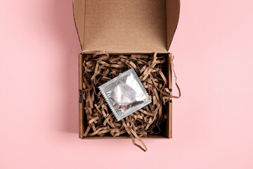 Open brown box with package of condom in brown shredded paper on pink background.