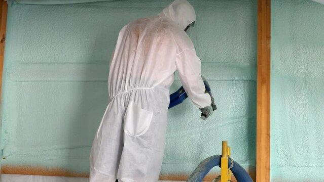 Worker in respirator and Tyvek suit sprays second closed cell foam insulation on the house wrap and studs of newly constructed exterior wall; concepts of hazards, protection and new construction