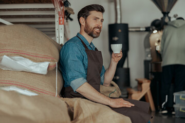 Business owner with cup of fresh coffee sitting on warehouse of coffee factory