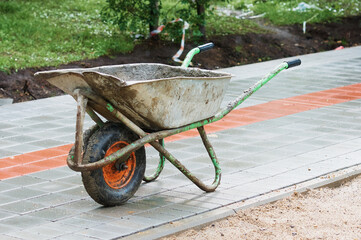 An old dirty metal wheelbarrow with an orange wheel on the sidewalk. Used construction tool, side view. Agricultural Wheel Farm Cart Flat Lawns Garden Cart Wheel Hand Cart