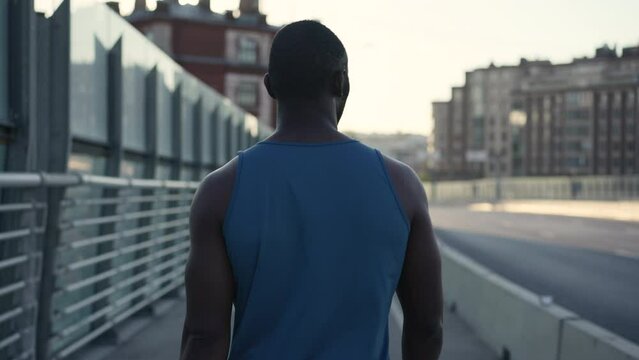 4k Back view of male athlete is walking on bridge during morning training in city spbas. Close view of American African man walks down street and watches urban views, poses for camera and rests from