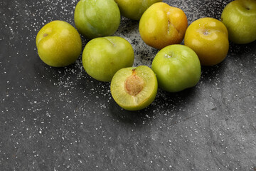 Small mini green fresh juicy plum fruit in wooden bowl on black slate stone board wooden table cut slice half seed sprinkled salt