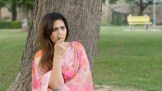 An attractive Indian sitting beside a tree and eating chocolate in a public park. A pretty female in casual clothes relaxing alone outdoors - sweet tooth