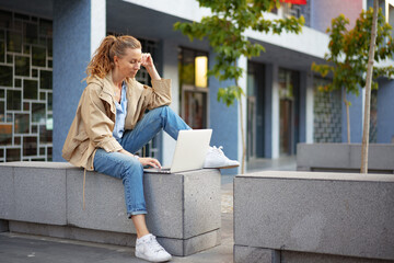 Fototapeta premium Young beautiful woman working on a laptop sitting on the bench in the street