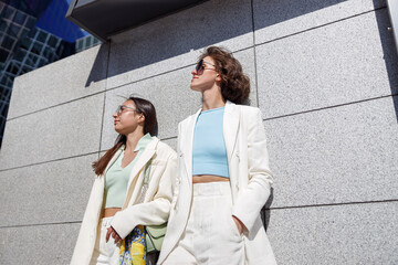 Fototapeta premium Two woman friends have a rest after walking in city standing near wall