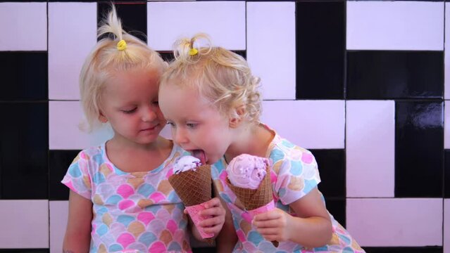 Two girls of three years old eat ice cream in waffle cones and share with each other. Two adorable twin girls eat ice cream together and try each other's tastes The pleasure of children from the sweet