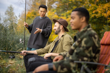 Asian man talking to male multiracial friends during fishing in nature. Men resting and spending...