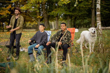 Fototapeta premium Pleased multiracial male friends resting and fishing on river or lake coast. Concept of leisure, hobby and weekend in nature. Idea of friendship and spending time together. View from the waterside