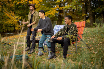 Pleased multiracial male friends resting and fishing on river or lake coast. Concept of leisure, hobby and weekend in nature. Idea of friendship and spending time together. View from the waterside
