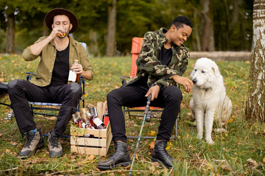 Two friends drinking strong alcohol sitting together with a dog on a lake coast near forest. Multiracial friends fishing and spending time together. Concept of leisure, hobby and weekend in nature