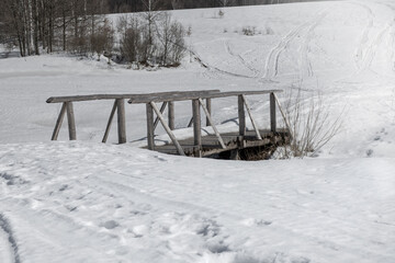 Winter landscape with a wooden bridge over a frozen stream and snow with trails in a hilly area in countryside.