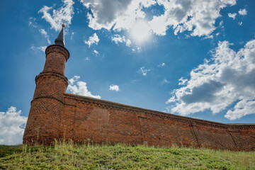 Part of a brick fortress wall with a tower against a blue cloudy sky and the sun.