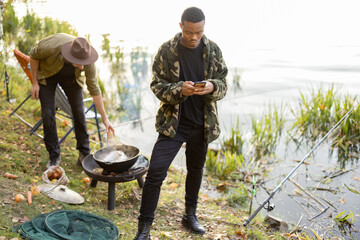 Hispanic men using smartphones during resting on river or pond coast with a friend cooking on background. Men fishing and cooking on river coast at autumn. Leisure and weekend in nature