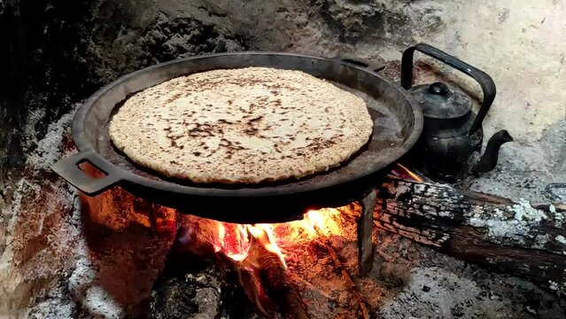 Close-up of Cooking bread in a cast iron skillet resting on trivets, over an oak wood fire in a large stone fireplace in a traditional kitchen. Medieval kitchen utensils. Morocco.